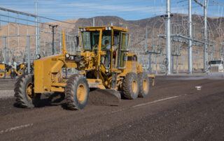Motor grader leveling gravel at a utility construction site with electrical infrastructure in the background and a drone capturing aerial footage.