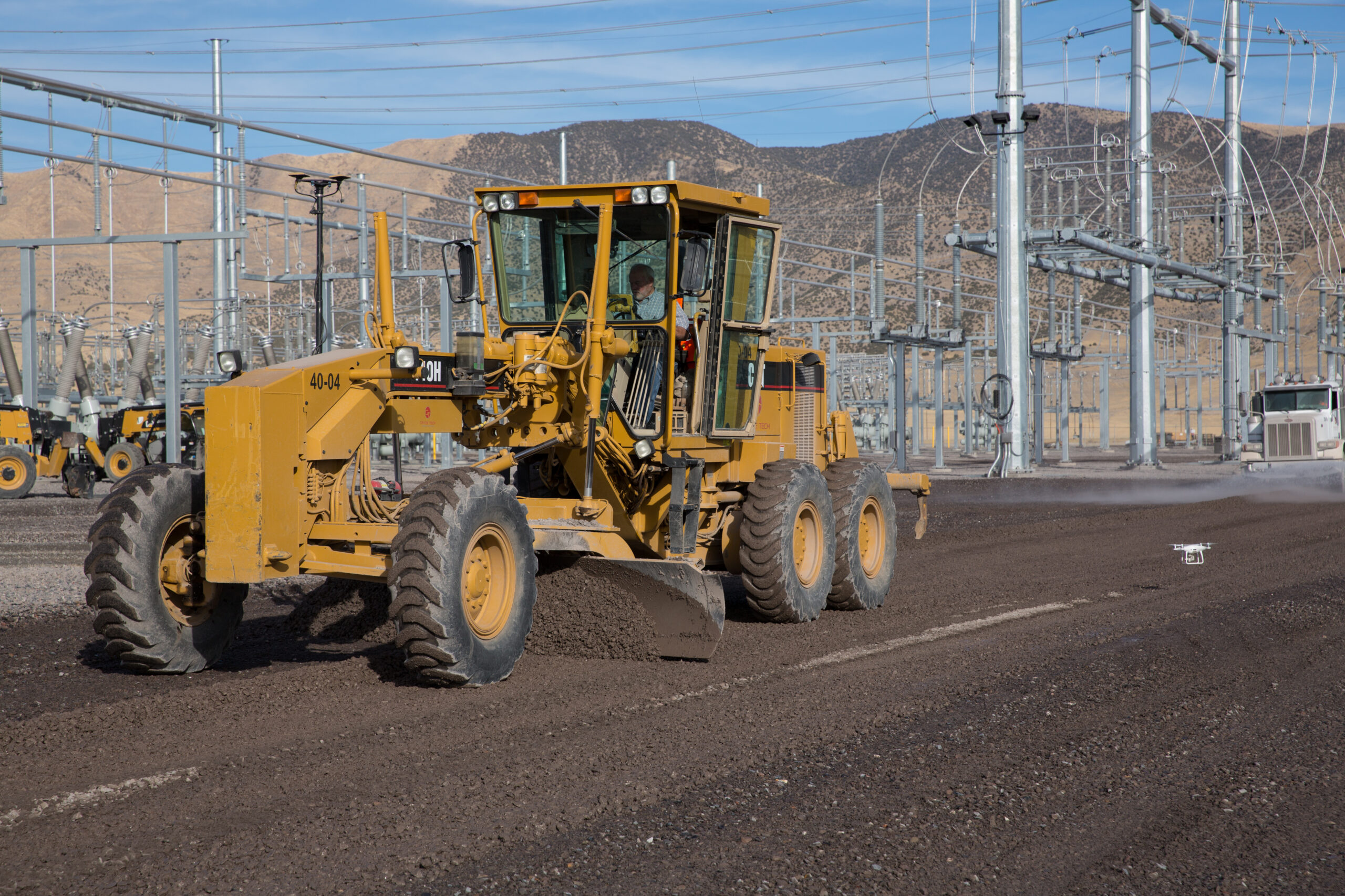 Motor grader leveling gravel at a utility construction site with electrical infrastructure in the background and a drone capturing aerial footage.