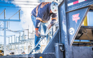 A worker in a hard hat and safety gear is securing a heavy-duty strap on a utility truck, likely part of equipment setup at a power substation. The background shows a complex arrangement of power lines and electrical components, suggesting an active work environment focused on energy infrastructure. The truck bears the logo of Grade Tech Power Services, indicating the worker is likely part of a team specializing in high-voltage or electrical construction projects.