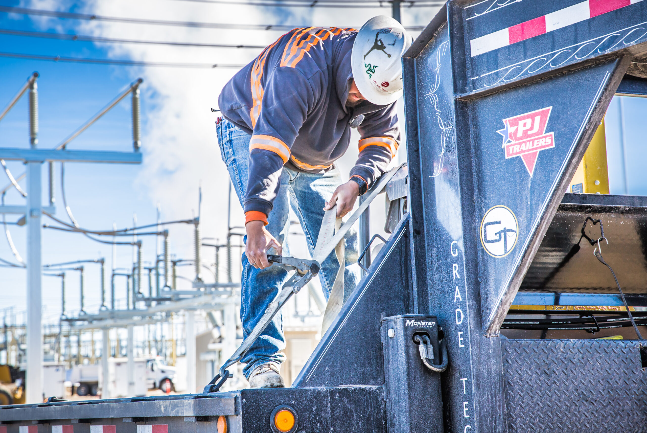 A worker in a hard hat and safety gear is securing a heavy-duty strap on a utility truck, likely part of equipment setup at a power substation. The background shows a complex arrangement of power lines and electrical components, suggesting an active work environment focused on energy infrastructure. The truck bears the logo of Grade Tech Power Services, indicating the worker is likely part of a team specializing in high-voltage or electrical construction projects.