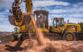 Construction site with two yellow Komatsu earthmoving machines—an excavator dumping dirt and a front loader—operated by workers wearing safety gear under a bright blue sky.
