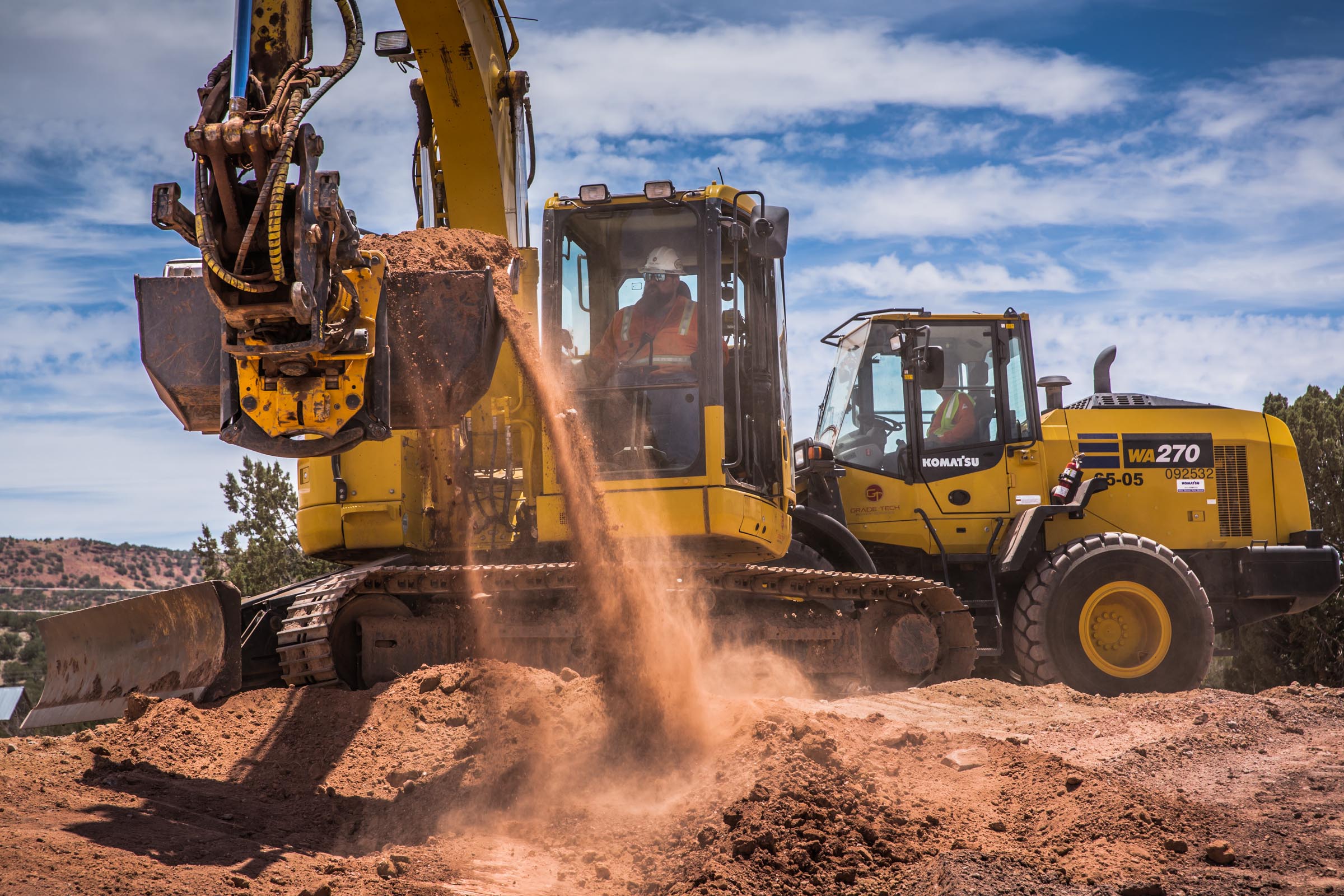 Construction site with two yellow Komatsu earthmoving machines—an excavator dumping dirt and a front loader—operated by workers wearing safety gear under a bright blue sky.