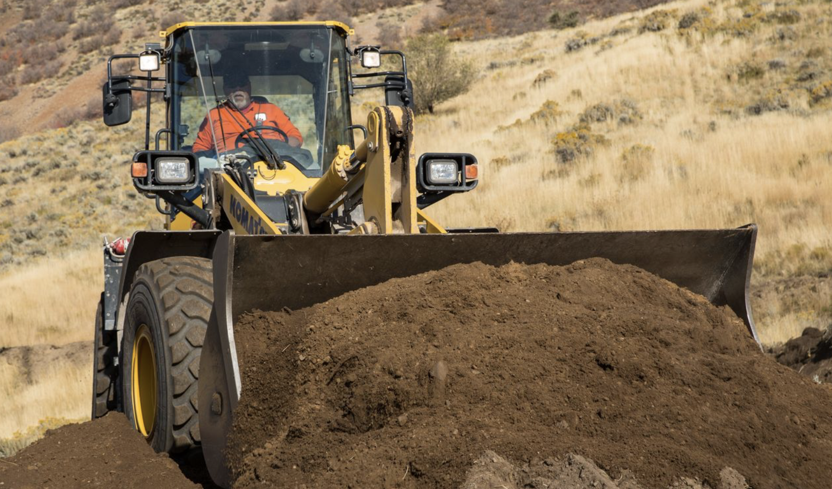 Construction worker operating a front-end loader moving soil at a job site in a dry hillside landscape.